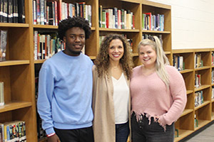 Three students smiling standing in the library in front of books.