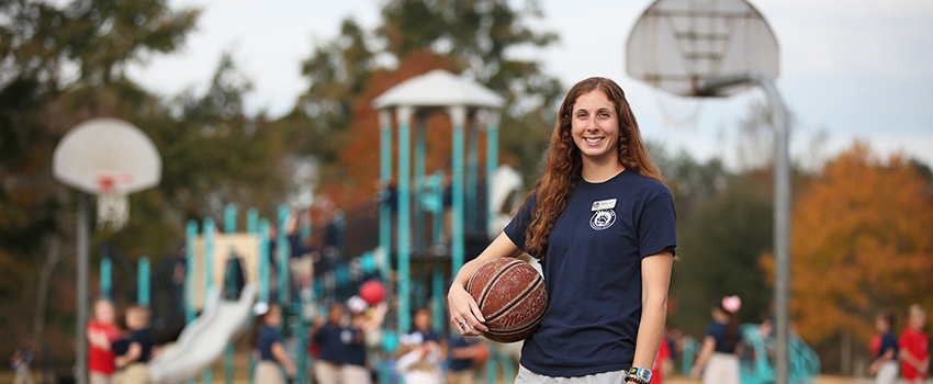 Female holding basketball in front of outdoor basketball court and playground.