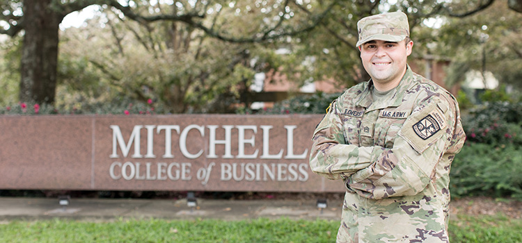 Ethan Flowers in uniform standing in front of Mitchell College of Business sign.