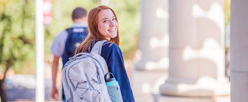 Student walking on campus smiling over shoulder with backpack.